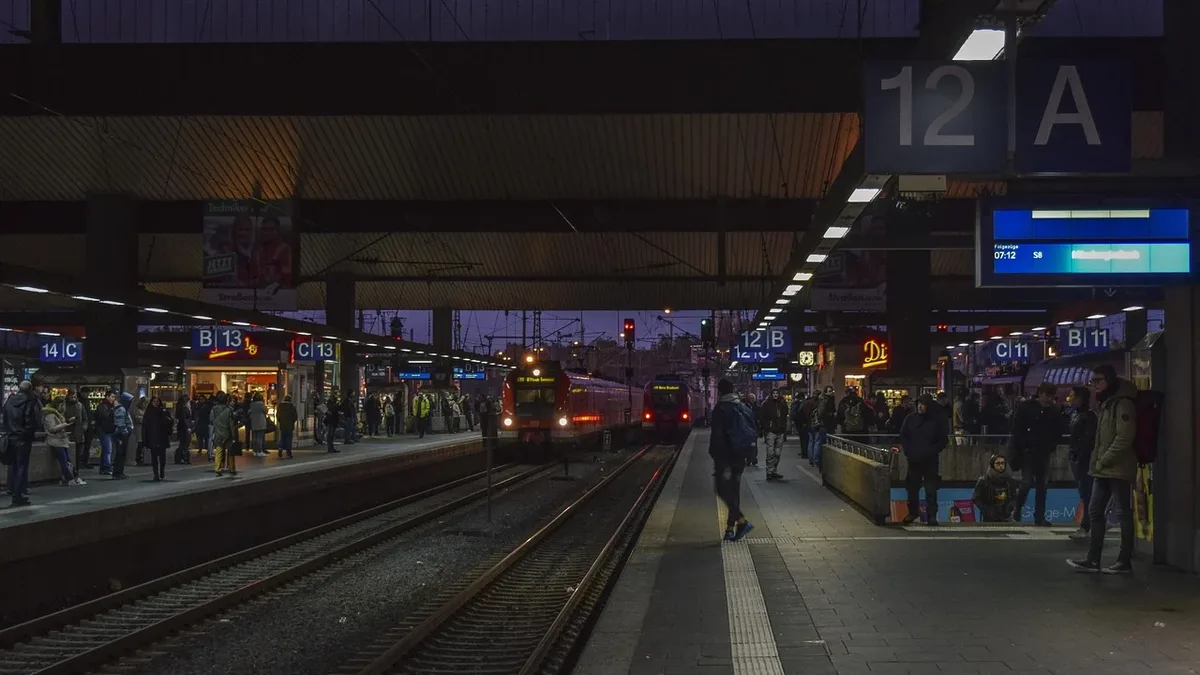 A train station with a train on the tracks, people waiting on the platform, and a sign indicating the platform number.